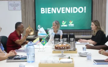Unesc e universidade colombiana discutem parcerias Reunião na Unesc, em Criciúma, com a reitora em exercício Gisele Silveira Coelho Lopes e o professor Alexis Calabari Angola da Universidad de La Guajira, Colômbia. Na mesa, pequenas bandeiras do Brasil e da Colômbia, garrafas de café e um bolo. Ao fundo, uma tela exibe a mensagem "BIENVENIDO!" com o logotipo da Unesc, marcando o início da visita institucional para parcerias acadêmicas e internacionalização.