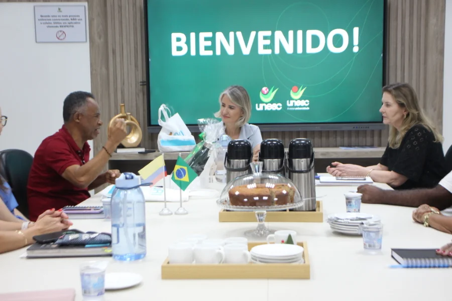 Reunião na Unesc, em Criciúma, com a reitora em exercício Gisele Silveira Coelho Lopes e o professor Alexis Calabari Angola da Universidad de La Guajira, Colômbia. Na mesa, pequenas bandeiras do Brasil e da Colômbia, garrafas de café e um bolo. Ao fundo, uma tela exibe a mensagem 