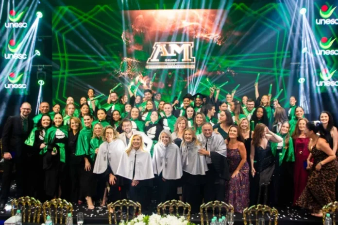 Unesc forma nova turma de Enfermagem em cerimônia especial Grupo de novos enfermeiros formados pela Unesc posa para foto oficial durante cerimônia de colação de grau, vestindo becas verdes e brancas em palco com iluminação festiva e logotipos da universidade.