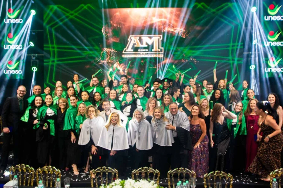 Unesc forma nova turma de Enfermagem em cerimônia especial Grupo de novos enfermeiros formados pela Unesc posa para foto oficial durante cerimônia de colação de grau, vestindo becas verdes e brancas em palco com iluminação festiva e logotipos da universidade.