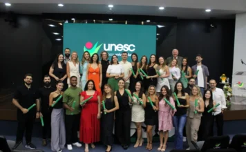 Unesc forma novos especialistas para o SUS em Criciúma Uma foto de grupo colorida, tirada no palco do Auditório Ruy Hülse, mostrando aproximadamente trinta novos especialistas das Residências Multiprofissionais da Unesc posando com seus canudos verdes de diploma. No fundo, um grande telão LED exibe o logotipo da 'unesc' em verde e a frase 'nossa universidade'. O grupo, vestido com roupas sociais variadas (vestidos coloridos, roupas pretas, camisas), está cercado por arranjos florais e um púlpito, com cadeiras de auditório vazias visíveis no primeiro plano. A imagem captura a celebração da conclusão do curso e a entrega de novos profissionais qualificados ao Sistema Único de Saúde (SUS) na região.