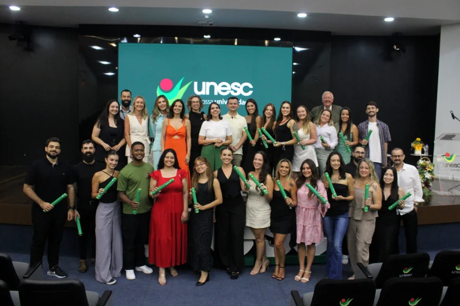 Uma foto de grupo colorida, tirada no palco do Auditório Ruy Hülse, mostrando aproximadamente trinta novos especialistas das Residências Multiprofissionais da Unesc posando com seus canudos verdes de diploma. No fundo, um grande telão LED exibe o logotipo da 'unesc' em verde e a frase 'nossa universidade'. O grupo, vestido com roupas sociais variadas (vestidos coloridos, roupas pretas, camisas), está cercado por arranjos florais e um púlpito, com cadeiras de auditório vazias visíveis no primeiro plano. A imagem captura a celebração da conclusão do curso e a entrega de novos profissionais qualificados ao Sistema Único de Saúde (SUS) na região.