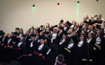 Unesc forma novos profissionais em dez cursos superiores Uma fotografia de médio alcance tirada durante a cerimônia de colação de grau da Unesc no Auditório Ruy Hülse. O palco está repleto de formandos de dez cursos, incluindo Adriano Ghellere, todos vestidos com becas pretas e capelos. Muitos seguram e levantam canudos de diplomas vermelhos e amarelos, com expressões de alegria e celebração. Alguns estão começando a tirar seus capelos e outros ajustam-nos. Luzes de palco coloridas (verde, vermelha, azul) iluminam o fundo. No primeiro plano, uma criança fotografa o momento com um celular.