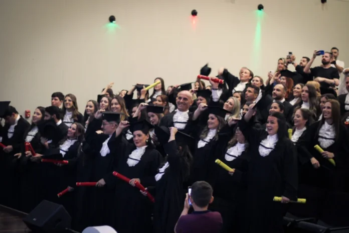 Unesc forma novos profissionais em dez cursos superiores Uma fotografia de médio alcance tirada durante a cerimônia de colação de grau da Unesc no Auditório Ruy Hülse. O palco está repleto de formandos de dez cursos, incluindo Adriano Ghellere, todos vestidos com becas pretas e capelos. Muitos seguram e levantam canudos de diplomas vermelhos e amarelos, com expressões de alegria e celebração. Alguns estão começando a tirar seus capelos e outros ajustam-nos. Luzes de palco coloridas (verde, vermelha, azul) iluminam o fundo. No primeiro plano, uma criança fotografa o momento com um celular.
