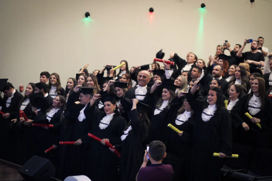 Uma fotografia de médio alcance tirada durante a cerimônia de colação de grau da Unesc no Auditório Ruy Hülse. O palco está repleto de formandos de dez cursos, incluindo Adriano Ghellere, todos vestidos com becas pretas e capelos. Muitos seguram e levantam canudos de diplomas vermelhos e amarelos, com expressões de alegria e celebração. Alguns estão começando a tirar seus capelos e outros ajustam-nos. Luzes de palco coloridas (verde, vermelha, azul) iluminam o fundo. No primeiro plano, uma criança fotografa o momento com um celular.