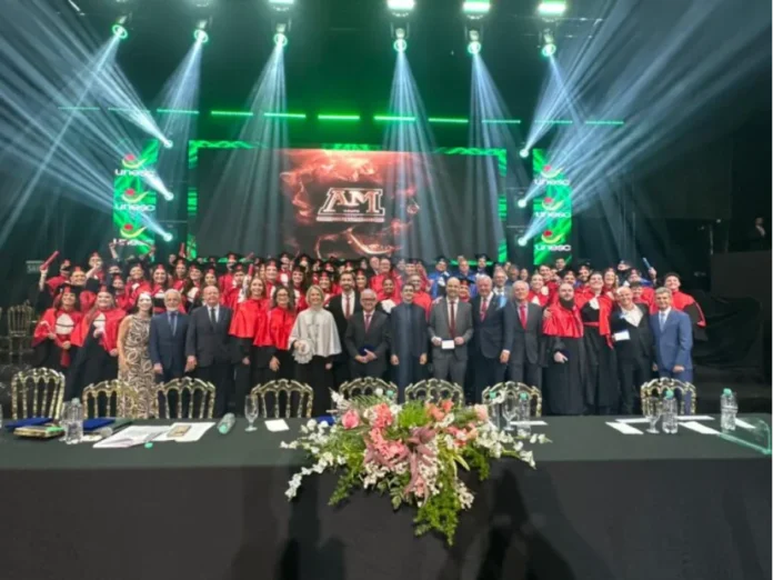 Unesc forma novos profissionais em noite de celebração Foto de grupo de formandos da UNESC com becas vermelhas e corpo docente em trajes formais no palco do AM Master Hall durante cerimônia de colação de grau em Criciúma. Luzes cênicas verdes e brancas no fundo com o logotipo da universidade e a inscrição "AM" na tela.