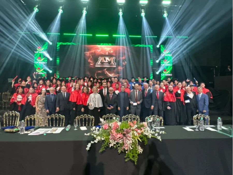 Foto de grupo de formandos da UNESC com becas vermelhas e corpo docente em trajes formais no palco do AM Master Hall durante cerimônia de colação de grau em Criciúma. Luzes cênicas verdes e brancas no fundo com o logotipo da universidade e a inscrição 