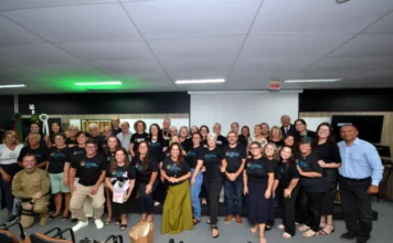 Unesc forma segunda turma de lideranças em Araranguá Grande foto de grupo capturada de frente, mostrando dezenas de pessoas vestindo camisetas pretas com a logomarca 'Lideranças Comunitárias Unesc' em um palco de auditório. No centro, os formandos e autoridades sorriem para a câmera. Inclui um homem de farda de bombeiro/policial à esquerda e o diretor do campus, Dorvanil Vieira, à direita, ambos com roupas formais. A imagem documenta a cerimônia de formatura da segunda turma da Escola de Lideranças Comunitárias da Unesc Araranguá, realizada em 25 de março de 2026. O auditório está com cadeiras vazias em primeiro plano e uma tela de projeção ao fundo.