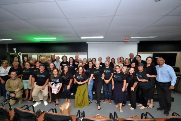 Unesc forma segunda turma de lideranças em Araranguá Grande foto de grupo capturada de frente, mostrando dezenas de pessoas vestindo camisetas pretas com a logomarca 'Lideranças Comunitárias Unesc' em um palco de auditório. No centro, os formandos e autoridades sorriem para a câmera. Inclui um homem de farda de bombeiro/policial à esquerda e o diretor do campus, Dorvanil Vieira, à direita, ambos com roupas formais. A imagem documenta a cerimônia de formatura da segunda turma da Escola de Lideranças Comunitárias da Unesc Araranguá, realizada em 25 de março de 2026. O auditório está com cadeiras vazias em primeiro plano e uma tela de projeção ao fundo.