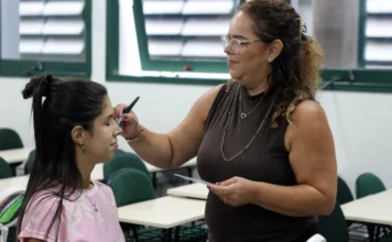 Unesc promove semana especial pelo Dia da Mulher Duas mulheres em uma sala de aula. À esquerda, uma jovem sentada com olhos fechados, cabelo escuro preso em um coque alto e vestindo uma camiseta rosa estampada. À direita, uma mulher madura de óculos com armação transparente, cabelos cacheados e blusa marrom sem mangas, está de pé e sorri enquanto aplica maquiagem no nariz da jovem com um pincel. A mulher madura segura um pequeno espelho de mão. Ao fundo, fileiras de carteiras escolares vazias e janelas de veneziana.