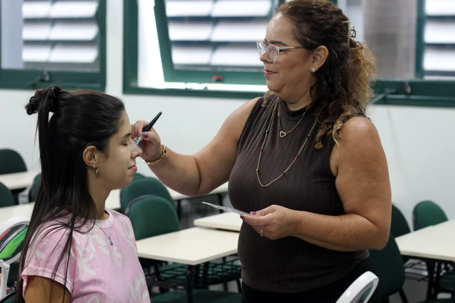 Duas mulheres em uma sala de aula. À esquerda, uma jovem sentada com olhos fechados, cabelo escuro preso em um coque alto e vestindo uma camiseta rosa estampada. À direita, uma mulher madura de óculos com armação transparente, cabelos cacheados e blusa marrom sem mangas, está de pé e sorri enquanto aplica maquiagem no nariz da jovem com um pincel. A mulher madura segura um pequeno espelho de mão. Ao fundo, fileiras de carteiras escolares vazias e janelas de veneziana.