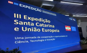Unesc recebe expedição da União Europeia em abril Grande tela de LED curvada em palco de auditório para a abertura da III Expedição Santa Catarina e União Europeia sobre fomento em ciência, tecnologia e inovação, um evento promovido pela Fapesc. Bandeiras de SC e da UE, e logotipos são visíveis. Arranjos de flores brancas no primeiro plano. A foto é de arquivo.