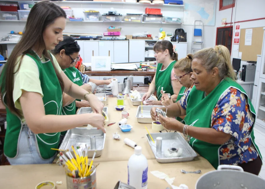 Grupo de mulheres participantes da oficina de velas artesanais da Unesc em uma sala de aula. Elas vestem aventais verdes com o logotipo da universidade e estão concentradas manuseando moldes, pavios e materiais para a fabricação das velas sobre mesas de trabalho. O ambiente é organizado, com prateleiras ao fundo contendo materiais de artesanato.