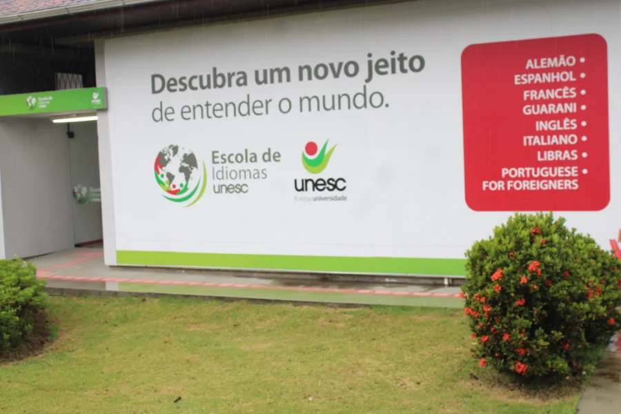 Unesc sedia evento inédito sobre ensino de idiomas Fachada da Escola de Idiomas da Unesc em Criciúma com painel exibindo a lista de cursos oferecidos, local que sedia o evento de formação para professores BRAZ-TESOL Santa Catarina.