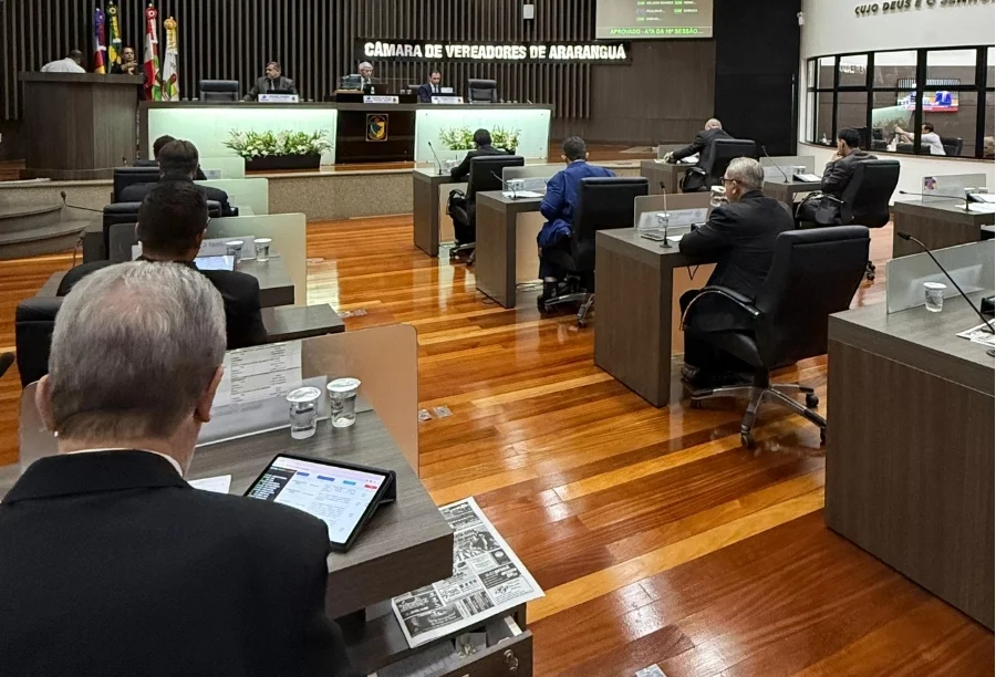 Sessão ordinária no plenário da Câmara de Vereadores de Araranguá com parlamentares sentados em suas bancadas durante votação de projetos de lei. A imagem mostra o ambiente legislativo com mesas de madeira, tablets e a mesa diretora ao fundo sob o letreiro da instituição.