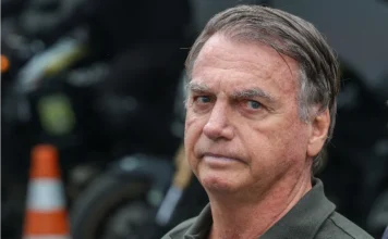 Defesa de Bolsonaro aguarda laudo para pedir domiciliar Jair Bolsonaro em primeiro plano com expressão séria e olhar fixo; ao fundo, motocicletas e sinalização de trânsito desfocadas durante evento público.