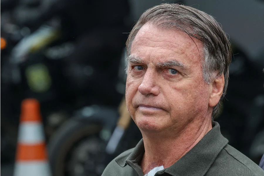 Jair Bolsonaro em primeiro plano com expressão séria e olhar fixo; ao fundo, motocicletas e sinalização de trânsito desfocadas durante evento público.