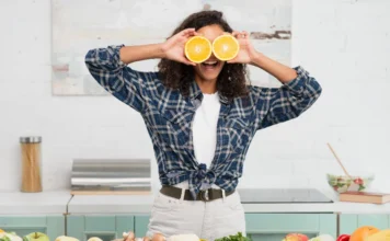 A influência da alimentação no bem-estar emocional! Mulher jovem e sorridente segura fatias de laranja nos olhos em uma cozinha repleta de vegetais frescos, ilustrando a conexão entre alimentação saudável, nutrição e bem-estar emocional.