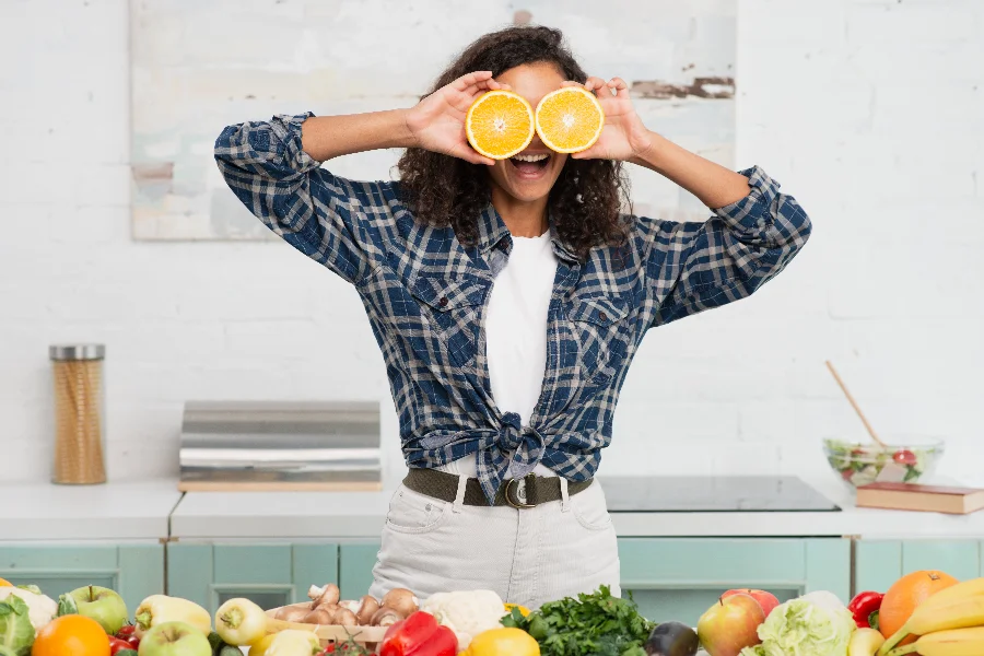 Mulher jovem e sorridente segura fatias de laranja nos olhos em uma cozinha repleta de vegetais frescos, ilustrando a conexão entre alimentação saudável, nutrição e bem-estar emocional.