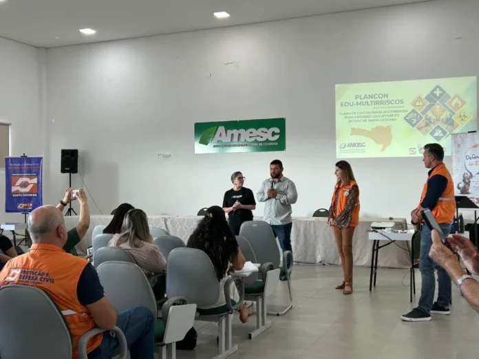 AMESC debate segurança escolar e prevenção de riscos em SC Representantes da AMESC e Defesa Civil de Santa Catarina em palestra sobre segurança escolar e o plano Plancon Edu-Multirriscos, com projeção e público presente em auditório.