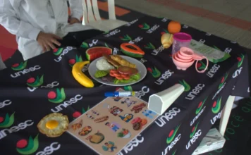 Acadêmicos da Unesc promovem nutrição descomplicada Acadêmicos do curso de Nutrição da Unesc apresentam réplicas de alimentos saudáveis e materiais educativos sobre uma mesa com o logotipo da universidade durante ação comunitária no hall da Reitoria.