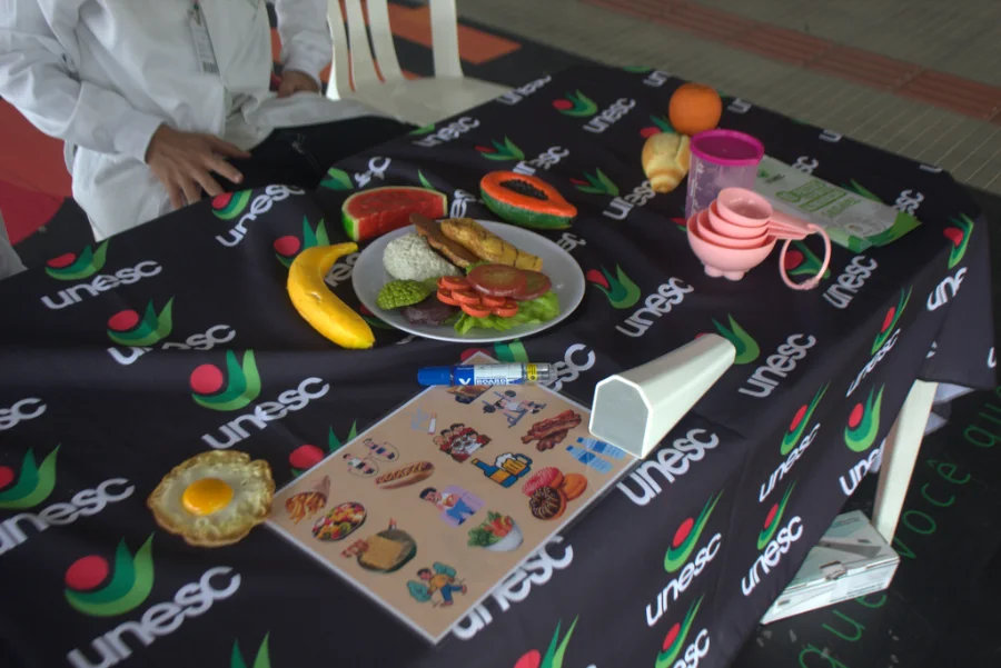 Acadêmicos do curso de Nutrição da Unesc apresentam réplicas de alimentos saudáveis e materiais educativos sobre uma mesa com o logotipo da universidade durante ação comunitária no hall da Reitoria.