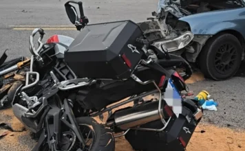 Acidente na SC-446 deixa um morto em Lauro Müller Cena de acidente de trânsito grave com colisão frontal entre uma motocicleta de aventura preta com malas laterais e um carro sedã azul claro na estrada. A motocicleta está caída no asfalto e a frente do carro está completamente destruída. Detritos e material absorvente de óleo estão espalhados na pista.
