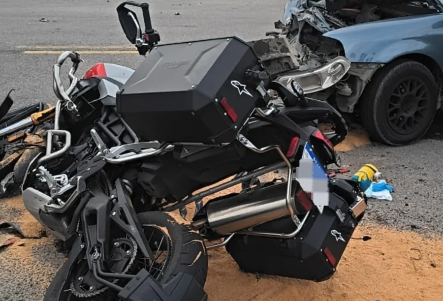Cena de acidente de trânsito grave com colisão frontal entre uma motocicleta de aventura preta com malas laterais e um carro sedã azul claro na estrada. A motocicleta está caída no asfalto e a frente do carro está completamente destruída. Detritos e material absorvente de óleo estão espalhados na pista.