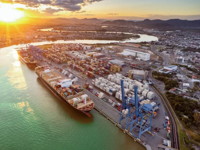 Acordo Mercosul-UE FIESC projeta novo ciclo para indústria Vista aérea de um terminal portuário em Santa Catarina com grandes navios cargueiros e milhares de contêineres ao pôr do sol, ilustrando a logística de exportação e o comércio internacional.