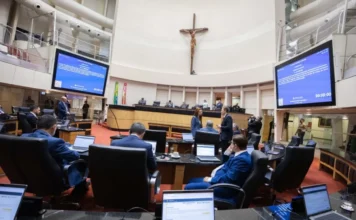 Alesc aprova presença de pais em consultas de menores Deputados estaduais em sessão no Plenário da Alesc, em Florianópolis, durante a votação do projeto de lei que garante a presença de ambos os pais em consultas médicas de menores. A imagem mostra os parlamentares em suas bancadas com notebooks e os painéis eletrônicos ao fundo.