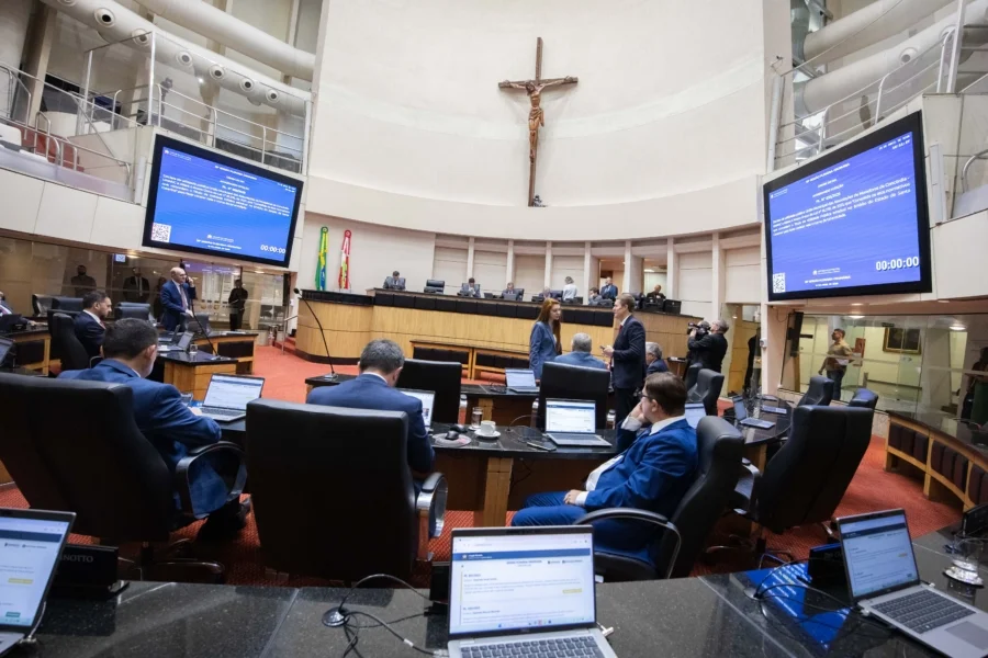 Deputados estaduais em sessão no Plenário da Alesc, em Florianópolis, durante a votação do projeto de lei que garante a presença de ambos os pais em consultas médicas de menores. A imagem mostra os parlamentares em suas bancadas com notebooks e os painéis eletrônicos ao fundo.
