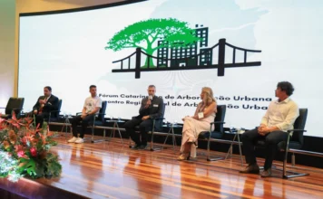 Alesc sedia fórum sobre arborização e clima em SC Uma foto panorâmica de um painel de discussão em um palco com piso de madeira. Cinco palestrantes estão sentados em cadeiras de escritório pretas. No centro, um homem de terno cinza fala ao microfone. À sua direita, uma mulher de vestido longo rosa claro. No fundo, um grande telão curvado exibe o logotipo do V Fórum Catarinense de Arborização Urbana, que apresenta uma grande árvore verde sobre uma ponte preta e silhuetas de prédios. O texto do evento está visível. Um arranjo de flores tropicais vermelhas está no canto inferior esquerdo do palco.