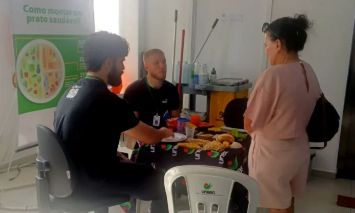 Alunos da Unesc realizam avaliação nutricional em Criciúma Acadêmicos de Nutrição da Unesc realizam ação de educação alimentar na UBS Centro de Criciúma, orientando paciente sobre como montar um prato saudável com modelos de alimentos sobre a mesa.