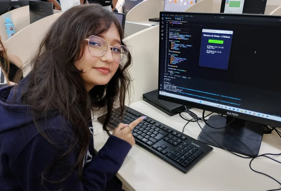 Estudante Eduarda Antara Cardoso aponta para tela de computador com códigos de programação do aplicativo de rotas de ônibus desenvolvido no curso técnico em Araranguá.