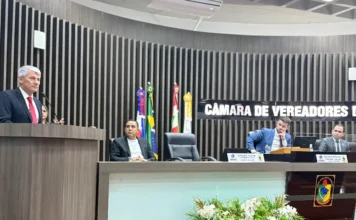 Araranguá: Projeto propõe fundo para desastres climáticos O vereador Paulinho Souza, do município de Araranguá, discursa na tribuna da Câmara de Vereadores, vestindo terno preto e gravata vermelha. A foto, capturada durante uma sessão ordinária, mostra o palestrante à esquerda e a mesa diretora ao fundo com outros parlamentares e bandeiras oficiais, retratando o momento da apresentação do projeto de lei para o fundo de defesa civil para desastres climáticos.