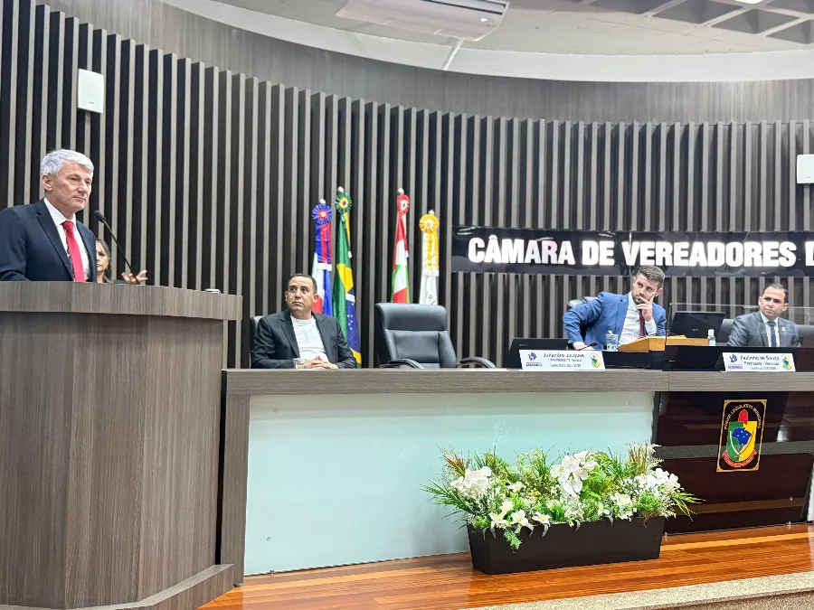 Araranguá Projeto propõe fundo para desastres climáticos O vereador Paulinho Souza, do município de Araranguá, discursa na tribuna da Câmara de Vereadores, vestindo terno preto e gravata vermelha. A foto, capturada durante uma sessão ordinária, mostra o palestrante à esquerda e a mesa diretora ao fundo com outros parlamentares e bandeiras oficiais, retratando o momento da apresentação do projeto de lei para o fundo de defesa civil para desastres climáticos.
