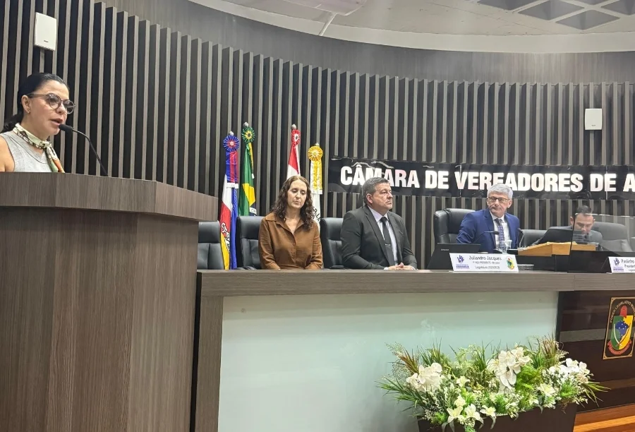 Coordenadora do Polo UAB de Araranguá, Maeve Espíndola Pereira, discursa na tribuna da Câmara de Vereadores sobre a abertura de inscrições para o curso gratuito de Administração Pública. A imagem detalha a palestrante em primeiro plano e a mesa diretora do legislativo ao fundo durante a 18ª sessão ordinária.