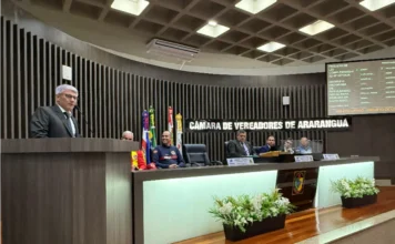 Araranguá aprova Refis Social com descontos de até 100% Presidente da Câmara de Araranguá, Paulinho Souza, discursando na tribuna sobre o Projeto de Lei do Refis Social para baixa renda. Placa da Câmara e tela com dados do PLC visíveis.