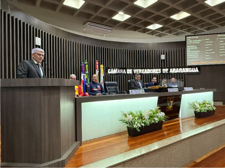 Presidente da Câmara de Araranguá, Paulinho Souza, discursando na tribuna sobre o Projeto de Lei do Refis Social para baixa renda. Placa da Câmara e tela com dados do PLC visíveis.