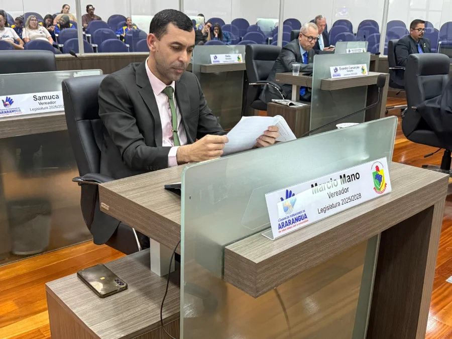 O vereador Márcio Mano sentado em sua bancada na Câmara Municipal de Araranguá, lendo o documento do anteprojeto de lei dos Jogos da Terceira Idade durante sessão ordinária. Ao fundo, outros vereadores e a galeria do plenário.