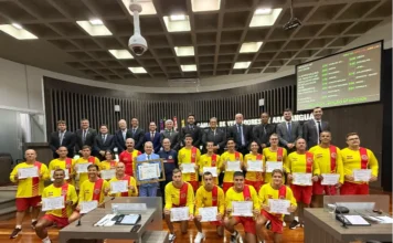 Araranguá aprova moção de reconhecimento a guarda-vidas Guarda-vidas civis e militares seguram certificados de Moção de Reconhecimento no plenário da Câmara de Vereadores de Araranguá, posando ao lado de vereadores e autoridades do Corpo de Bombeiros pela atuação na Operação Veraneio 2026 no Morro dos Conventos.