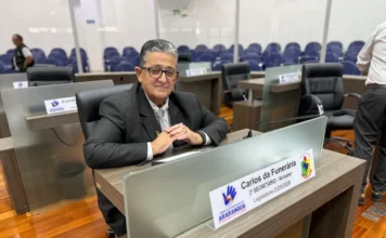 Araranguá aprova projeto da ‘Creche do Idoso’ na Câmara Vereador Carlos da Funerária, de terno e óculos, sentado em sua bancada no plenário da Câmara de Vereadores de Araranguá. Ele sorri para a foto, tendo ao fundo as cadeiras azuis do auditório e uma placa de identificação da 2ª Secretaria da Legislatura 2025/2028.