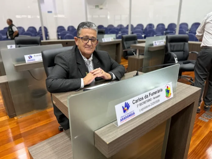 Araranguá aprova projeto da 'Creche do Idoso' na Câmara Vereador Carlos da Funerária, de terno e óculos, sentado em sua bancada no plenário da Câmara de Vereadores de Araranguá. Ele sorri para a foto, tendo ao fundo as cadeiras azuis do auditório e uma placa de identificação da 2ª Secretaria da Legislatura 2025/2028.