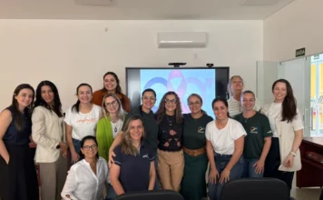 Araranguá capacita enfermeiros para testes de HIV e sífilis Grupo de enfermeiras da rede municipal de saúde de Araranguá reunidas em sala de treinamento durante capacitação técnica sobre tuberculose e testes rápidos de HIV e sífilis. Ao fundo, uma tela exibe símbolos de conscientização em saúde.