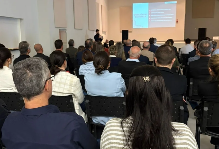 Público de costas em auditório acompanhando palestra técnica sobre os impactos da Reforma Tributária em Araranguá. Ao fundo, palestrante próximo a tela de projeção exibindo slide com o título Ineficiências Cadastrais para gestores e servidores públicos.