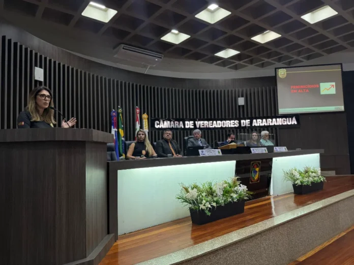 Araranguá discute estratégias de combate ao feminicídio Delegada Eliane Chaves discursa na tribuna da Câmara de Vereadores de Araranguá sobre o aumento dos casos de feminicídio em Santa Catarina. Ao fundo, mesa diretora da casa e tela de projeção com o alerta "Feminicídios em alta".