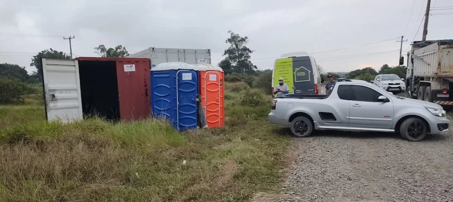 Canteiro de obras inicial em Araranguá para a 4ª ponte, mostrando contêineres, banheiros químicos azul e laranja, caminhonetes Chevrolet Montana e outros veículos da equipe de engenharia.