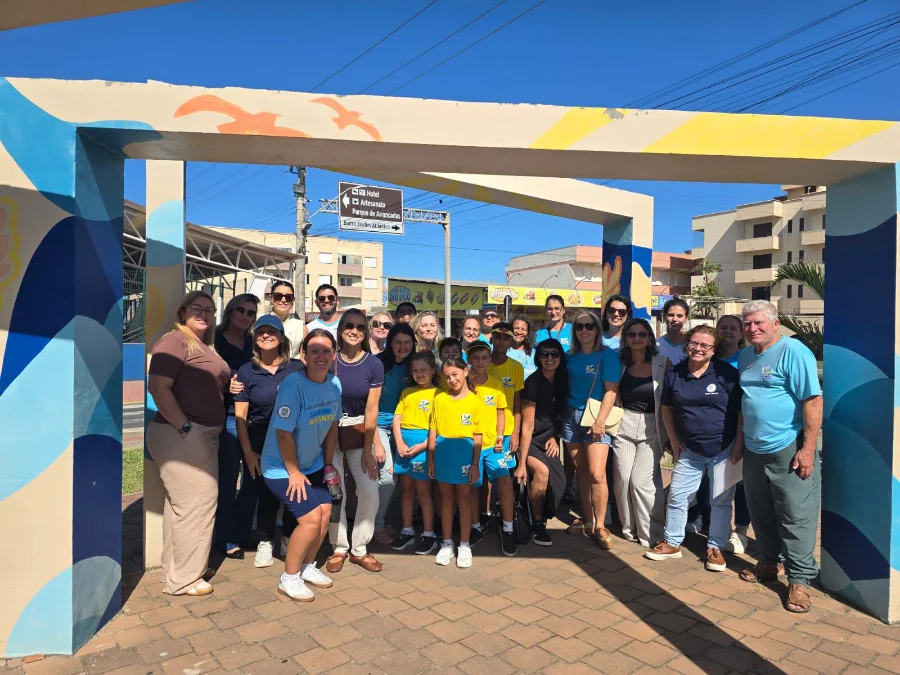 Grupo de pessoas, alunos e pais, posando para foto na Praça Central de Balneário Arroio do Silva durante o evento Dia da Família na Escola. As crianças e adultos vestem uniformes azuis e amarelos da Escola Adventista Jardim Atlântico. Eles estão emoldurados por um portal colorido com gaivotas. Placas turísticas e prédios são visíveis ao fundo sob céu limpo.