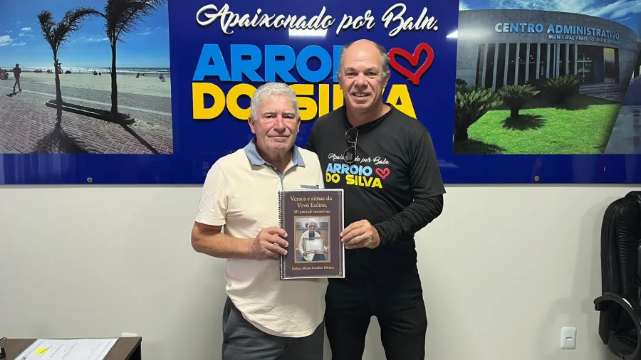O prefeito de Balneário Arroio do Silva, Jorge Luiz Freitas, à direita, recebe do neto de Dona Eulina, Everson (Xande), o livreto de versos em homenagem aos 101 anos da moradora centenária. Ambos seguram a obra literária em frente a um painel institucional do município.