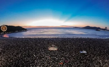 BC: 15 Mil pessoas oram ao amanhecer na Sexta-Feira Santa Vista aérea de multidão reunida na areia da Praia Central de Balneário Camboriú durante o evento Epiphany Movement ao amanhecer da Sexta-feira Santa. Ao centro, um letreiro iluminado escrito JESUS e a roda-gigante FG Big Wheel ao fundo à esquerda sob o céu do nascer do sol.